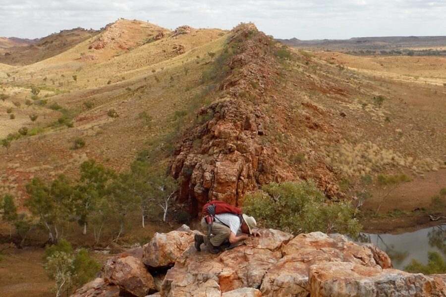 A man studies a rock on top of a hill with prairie-covered hills in the background.